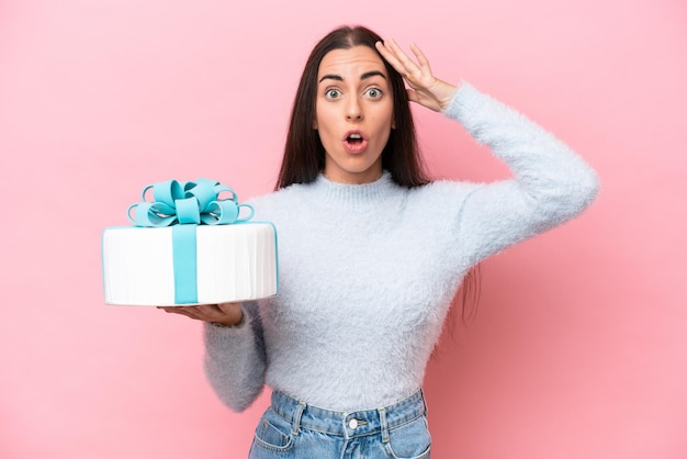 Young caucasian woman holding birthday cake isolated on pink background with surprise expression