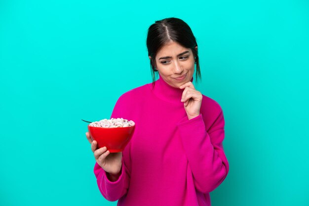 Young caucasian woman holding a bowl of cereals isolated on blue background with sad expression