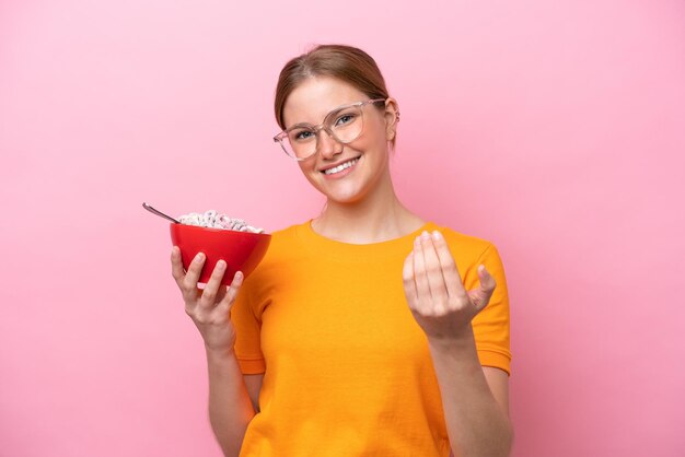 Photo young caucasian woman holding a bowl of cereals isolated on pink background inviting to come with hand happy that you came