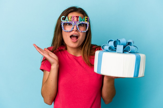 Young caucasian woman holding a cake isolated on blue background surprised and shocked.