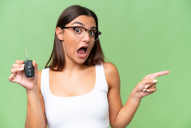 Young caucasian woman holding car keys isolated on green chroma background background surprised and pointing finger to the side