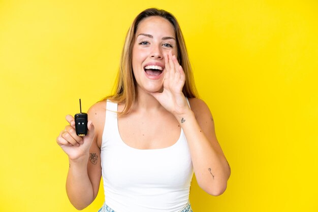 Young caucasian woman holding car keys isolated on yellow background shouting with mouth wide open