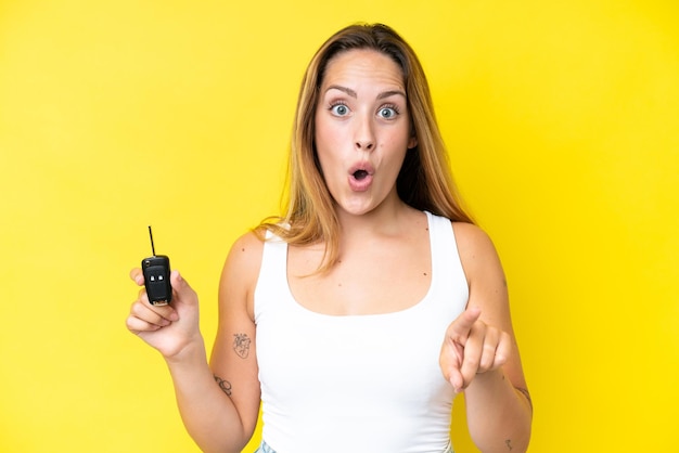 Young caucasian woman holding car keys isolated on yellow background surprised and pointing front