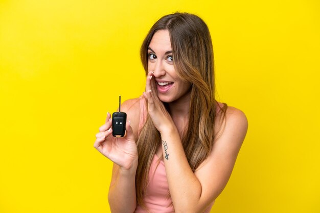 Young caucasian woman holding car keys isolated on yellow background whispering something