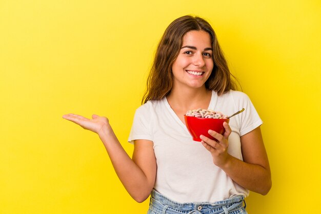 Young caucasian woman holding cereals isolated on yellow background  showing a copy space on a palm and holding another hand on waist.