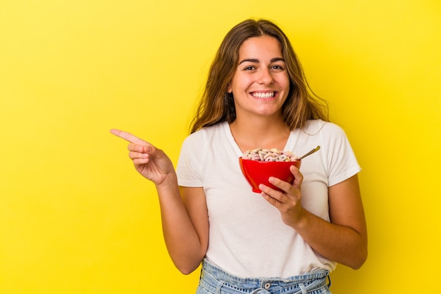 Young caucasian woman holding cereals isolated on yellow background  smiling and pointing aside, showing something at blank space.