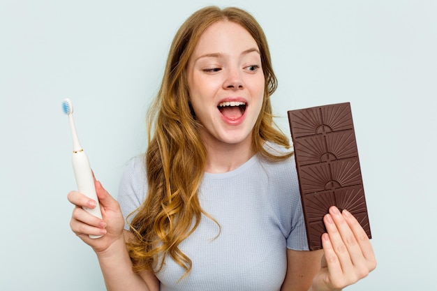 Young caucasian woman holding chocolate and toothbrush isolated on blue background