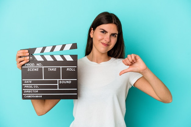 Young caucasian woman holding a clapperboard isolated on blue background feels proud and self confident, example to follow.