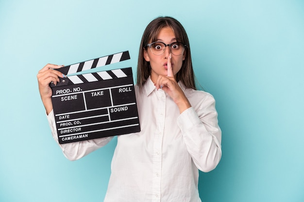 Young caucasian woman holding clapperboard isolated on blue background keeping a secret or asking for silence.