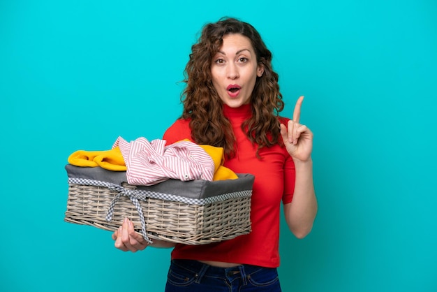 Young caucasian woman holding a clothes basket isolated on blue background intending to realizes the solution while lifting a finger up