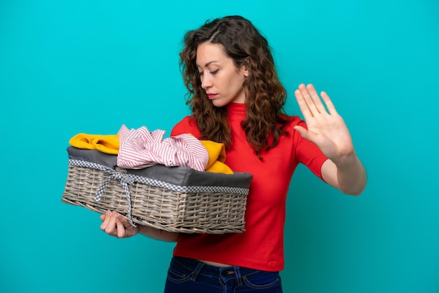 Young caucasian woman holding a clothes basket isolated on blue background making stop gesture and disappointed