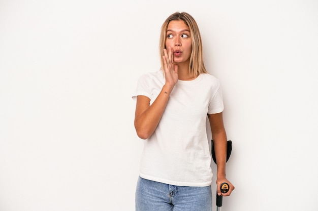 Young caucasian woman holding crutch isolated on white background is saying a secret hot braking news and looking aside