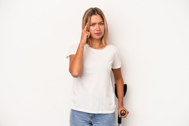 Young caucasian woman holding crutch isolated on white background pointing temple with finger, thinking, focused on a task.