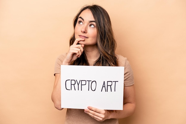 Young caucasian woman holding a crypto art placard isolated on beige background looking sideways with doubtful and skeptical expression