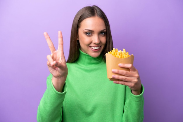 Young caucasian woman holding fried chips on purple background smiling and showing victory sign