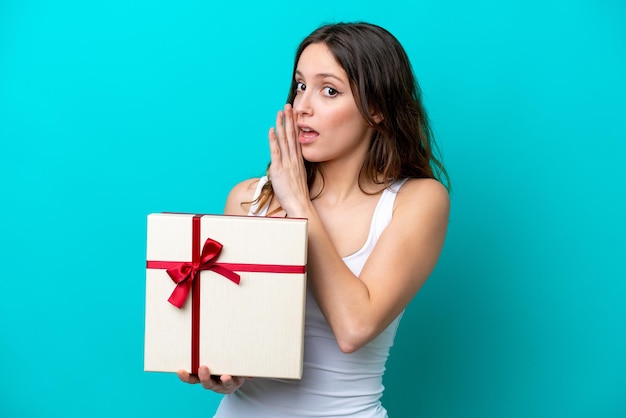 Young caucasian woman holding a gift isolated on blue background whispering something