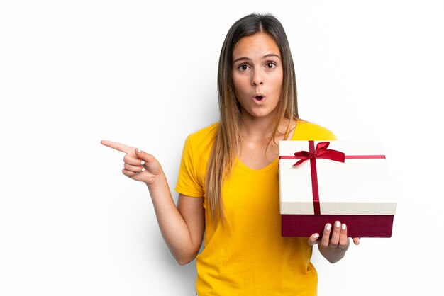 Young caucasian woman holding a gift isolated on white background surprised and pointing side