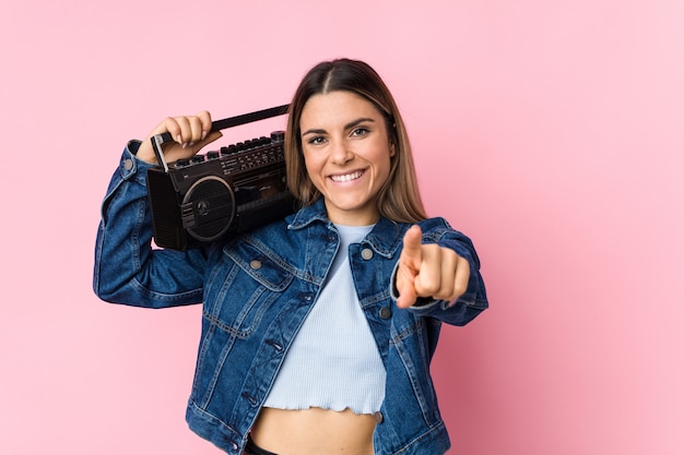 Young caucasian woman holding a guetto blaster cheerful smiles pointing to front.