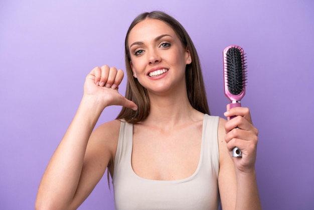 Young caucasian woman holding hairbrush isolated on purple background proud and selfsatisfied