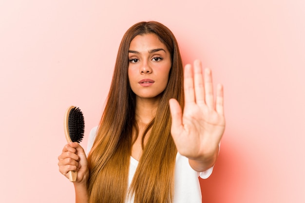 Young caucasian woman holding an hairbrush standing with outstretched hand showing stop sign, preventing you.