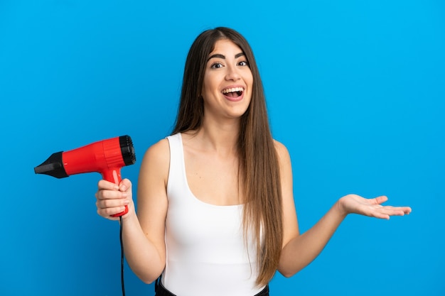 Young caucasian woman holding a hairdryer isolated on blue background with shocked facial expression