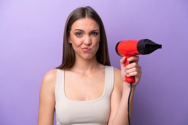 Young caucasian woman holding a hairdryer isolated on purple background with sad expression