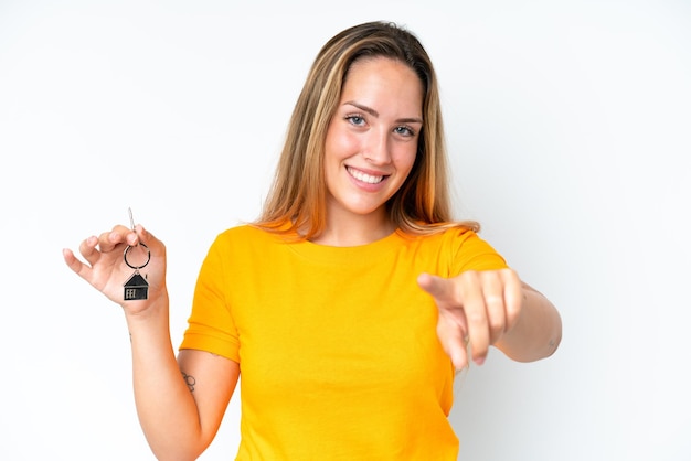 Young caucasian woman holding home keys isolated on white background points finger at you with a confident expression