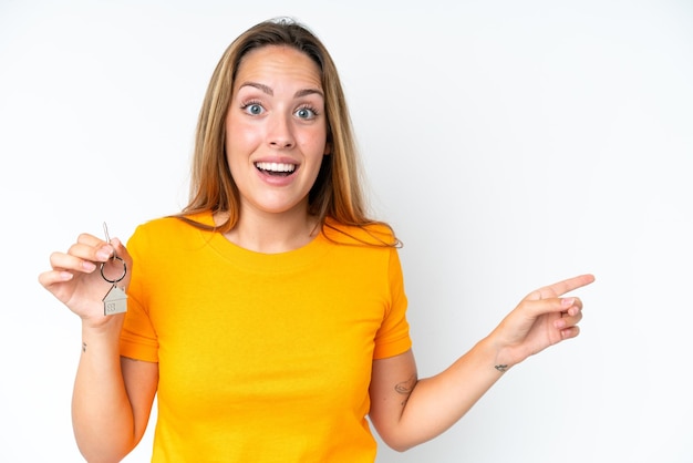 Young caucasian woman holding home keys isolated on white background surprised and pointing finger to the side