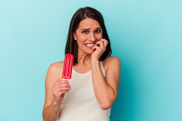 Young caucasian woman holding an ice cream isolated on blue background biting fingernails nervous and very anxious