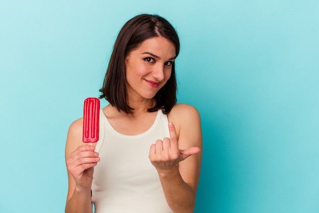 Young caucasian woman holding an ice cream isolated on blue background pointing with finger at you as if inviting come closer.