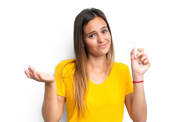 Young caucasian woman holding invisible braces isolated on white background making doubts gesture while lifting the shoulders