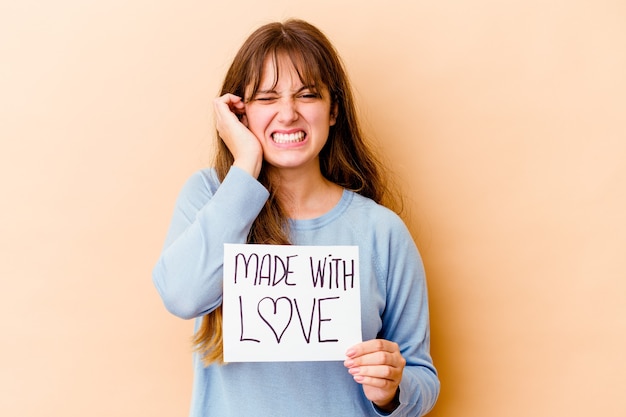 Young caucasian woman holding a made with love placard isolated covering ears with hands.