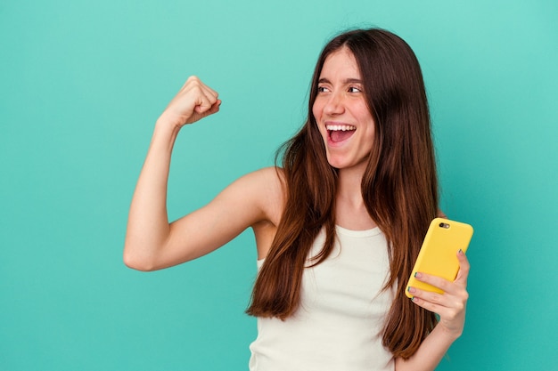 Young caucasian woman holding a mobile phone isolated on blue background raising fist after a victory, winner concept.