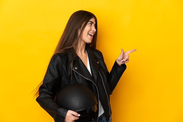 Young caucasian woman holding a motorcycle helmet isolated on yellow wall pointing finger to the side and presenting a product