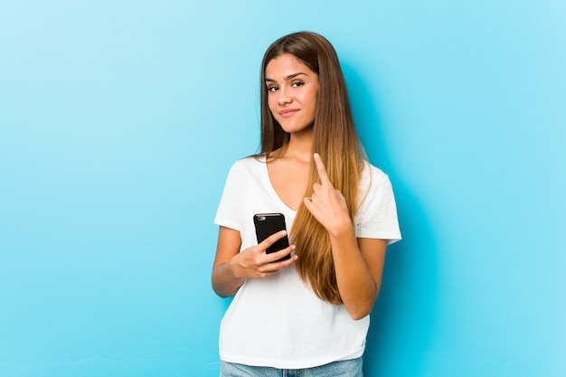 Young caucasian woman holding a phone pointing with finger at you as if inviting come closer.