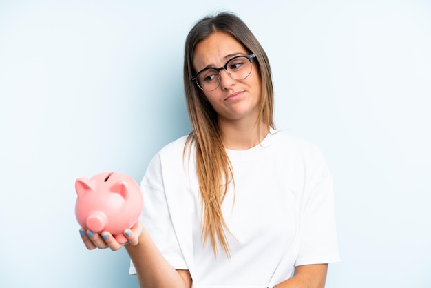 Young caucasian woman holding a piggybank isolated on blue background with sad expression