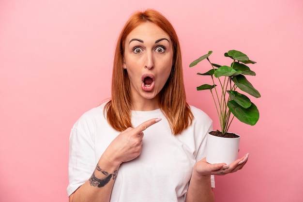 Young caucasian woman holding a plant isolated on pink background Young caucasian woman holding a plant isolated on pink background pointing to the side