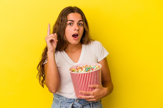 Young caucasian woman holding a popcorns isolated on yellow background  having an idea, inspiration concept.