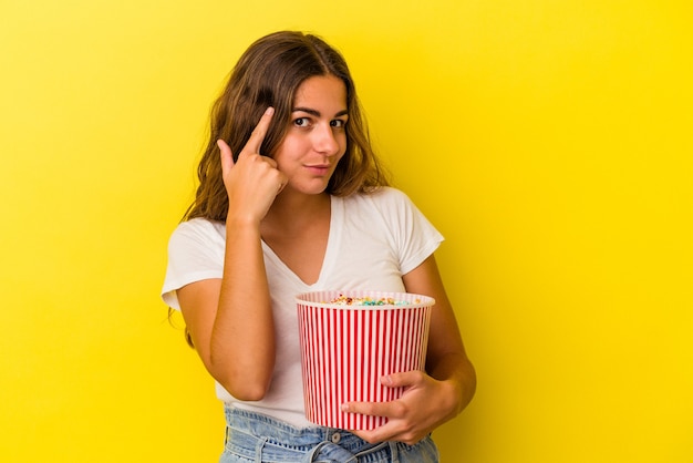 Young caucasian woman holding a popcorns isolated on yellow background  pointing temple with finger, thinking, focused on a task.