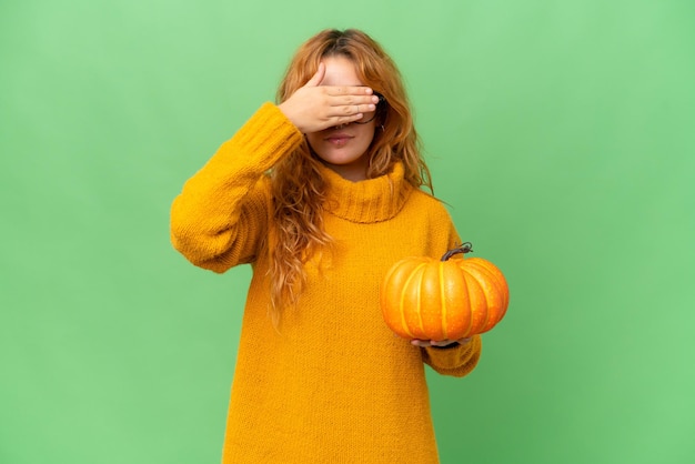 Young caucasian woman holding a pumpkin isolated on green screen chroma key background covering eyes by hands Do not want to see something