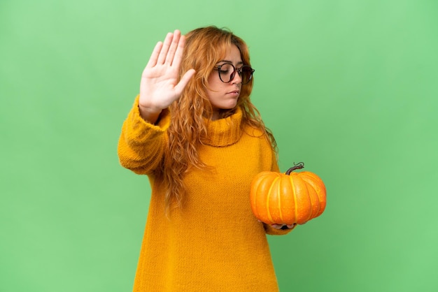 Young caucasian woman holding a pumpkin isolated on green screen chroma key background making stop gesture and disappointed