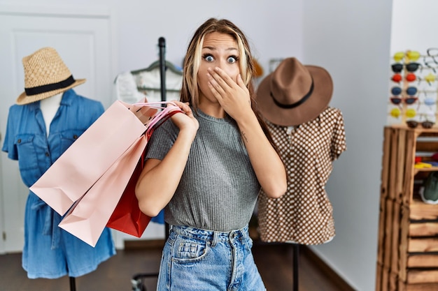 Young caucasian woman holding shopping bags at retail, shop covering mouth with hand, shocked and afraid for mistake. surprised expression