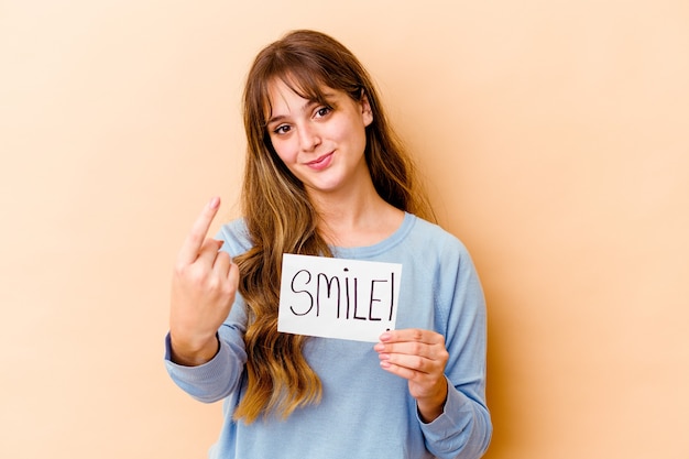 Young caucasian woman holding a Smile placard isolated pointing with finger at you as if inviting come closer.