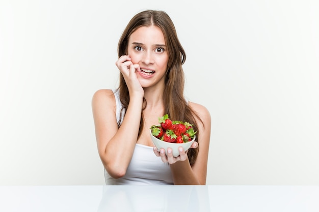 Young caucasian woman holding a strawberries bowl biting fingernails, nervous and very anxious.