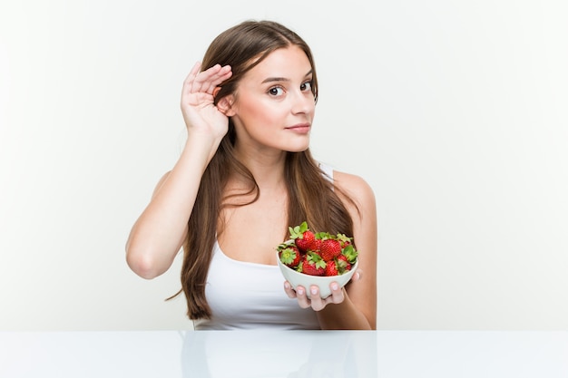 Young caucasian woman holding a strawberries bowl trying to listening a gossip.