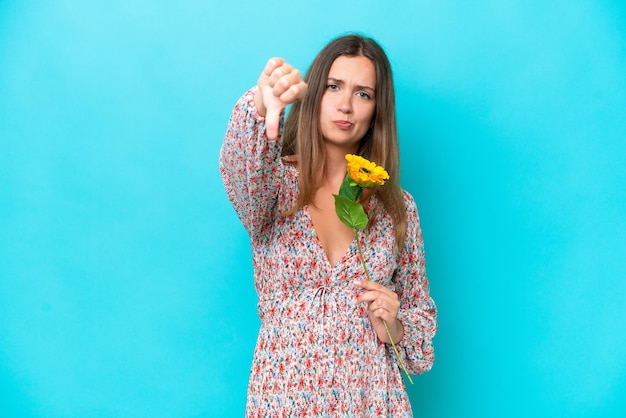 Young caucasian woman holding sunflower isolated on blue background showing thumb down with negative expression