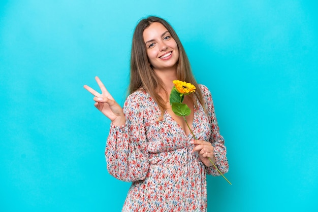 Young caucasian woman holding sunflower isolated on blue background smiling and showing victory sign