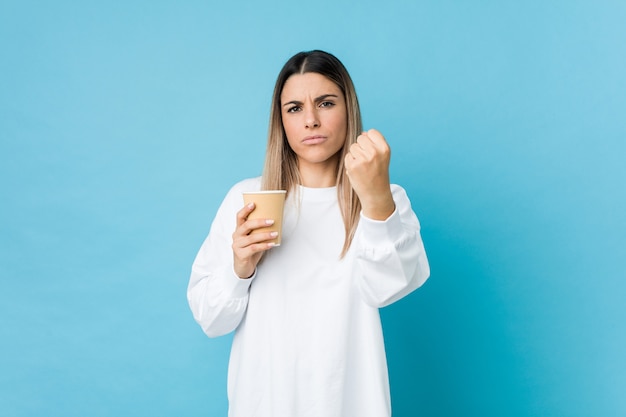 Young caucasian woman holding a takeaway coffee showing fist to camera, aggressive facial expression.