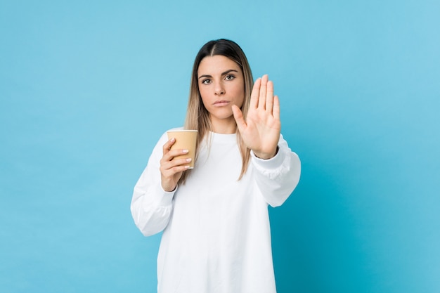 Young caucasian woman holding a takeaway coffee standing with outstretched hand showing stop sign, preventing you.