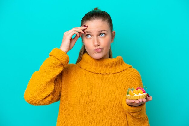 Young caucasian woman holding a tartlet isolated on blue background having doubts and with confuse face expression